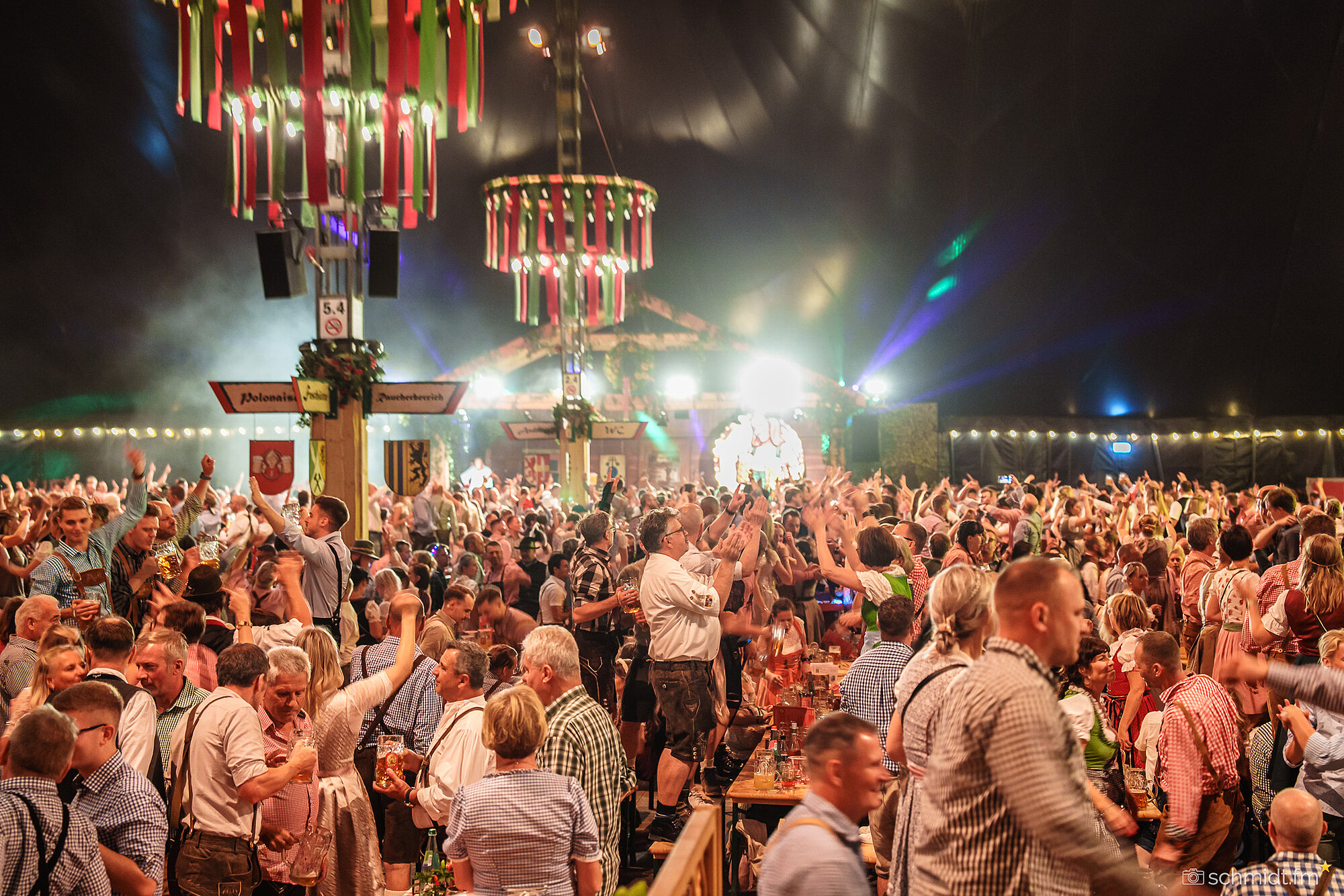 Festlich dekoriertes Bierzelt mit Menschen in Dirndl und Lederhosen, die auf langen Holzbänken stehen und feiern