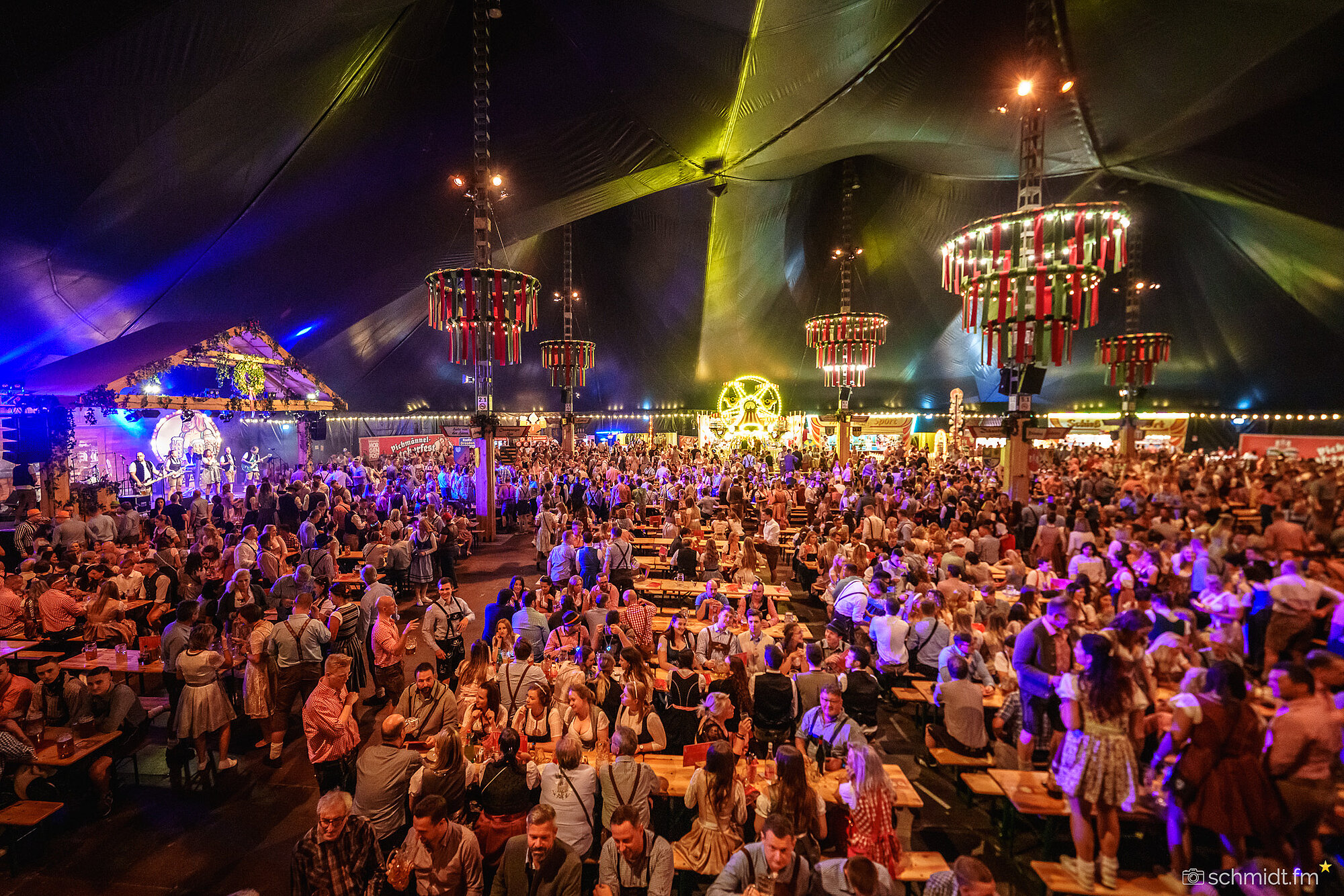 Festlich dekoriertes Bierzelt mit Menschen in Dirndl und Lederhosen, die an langen Holztischen sitzen und Bierkrüge in die Luft halten