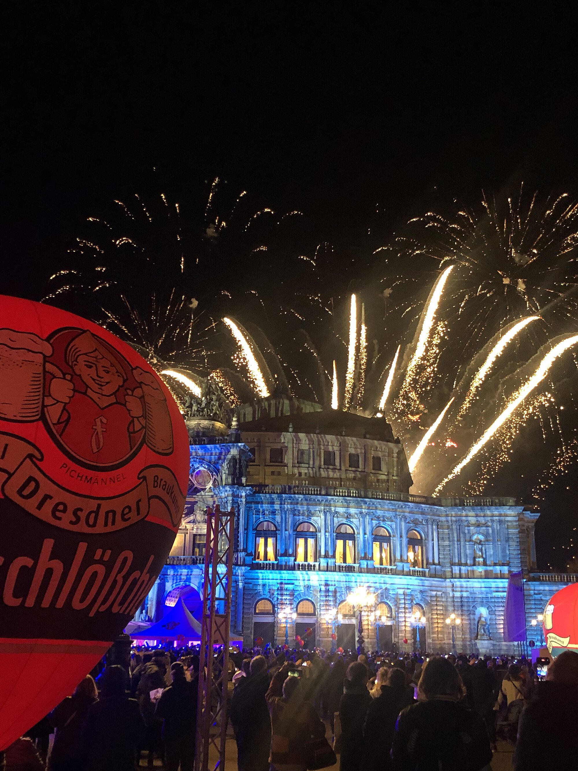 Eine Menschenmenge vor der blau beleuchtete Semperoper mit Feuerwerk im Hintergrund