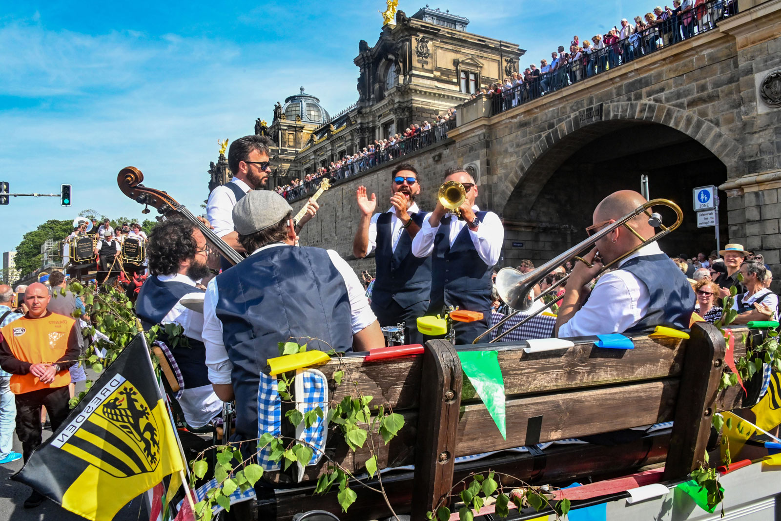 Die historische Dresdner Altstadt mit einer Jazzband im Zentrum und der Carolabrücke im Hintergrund. Die Band spielt und Zuschauer jubeln von der Brücke während Jazzklänge die Luft erfüllen.